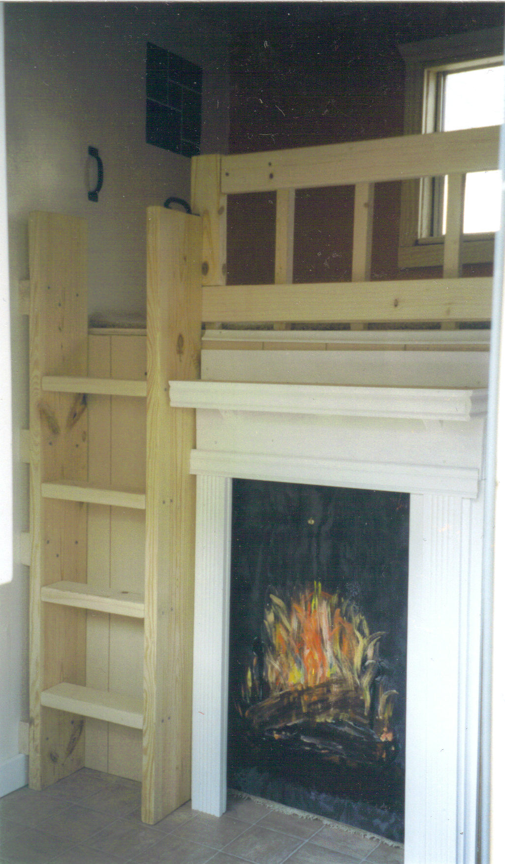 Inside the "birdhaus," photo of the ladder that leads to the loft. A fake, painted fireplace sits to the right of the ladder. Though this room is tiled, the space behind the fireplace appears to be carpeted. The loft has a window and a railing, and there are handles on the wall to assist with climbing. Blue-and-green rectangular wall art can be seen.