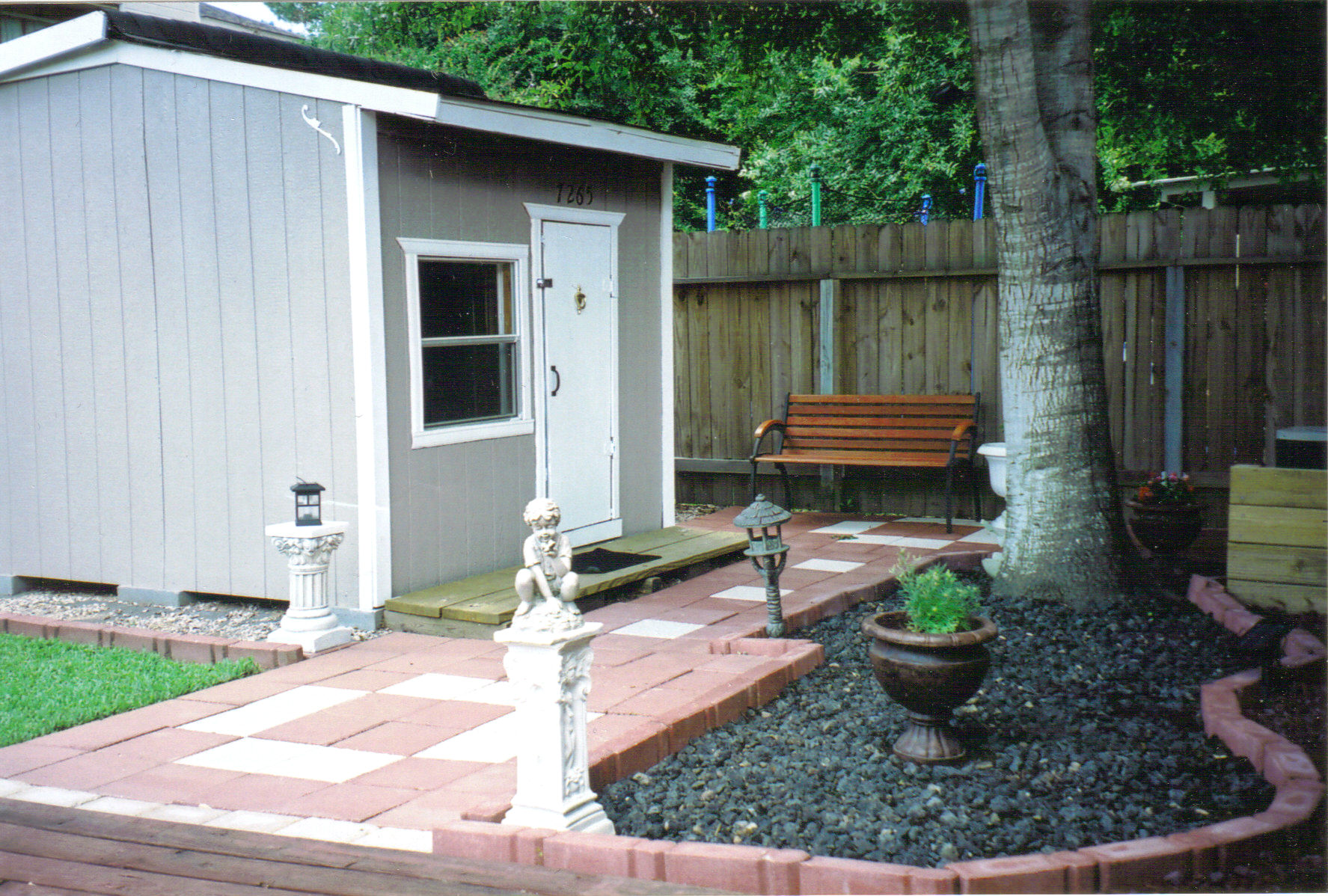 A "playhouse" in a cute backyard. It has grey siding, a window, a door, shingled roof, and even a street number.